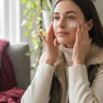 Young woman applying moisturizer on her face during winter, showing healthy glowing skin.